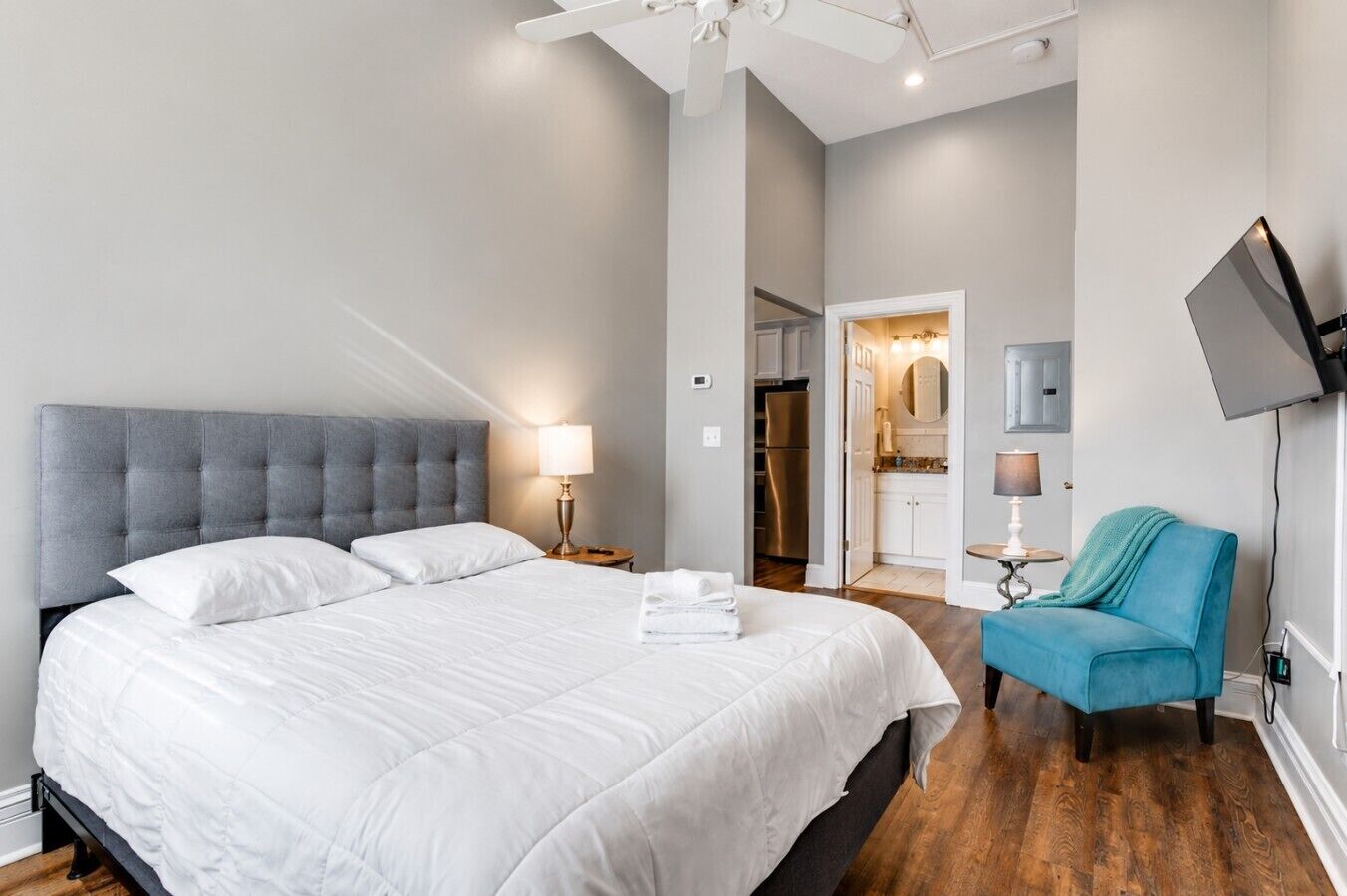 Un dormitorio con una cama grande, un cabecero gris, una lámpara de noche, un sillón azul y un televisor montado en la pared.