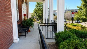 A covered porch with a brick wall, white columns, a bench, and a planter with flowers. - Bright & Cozy Church Conversion • 1BR (Nashville)