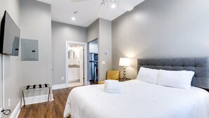 A bedroom with a large bed, a ceiling fan, a TV mounted on the wall, and a visible bathroom door. - Bright & Cozy Church Conversion • 1BR (Nashville)