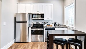A compact kitchen with stainless steel appliances, white cabinets, a dining area with a dark wood table and two chairs, and a window with blinds. - Bright & Cozy Church Conversion • 1BR (Nashville)