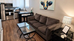 Un espace de vie compact comprenant un coin cuisine, un canapé marron, une table basse en verre et un tableau représentant une vache accroché au mur.