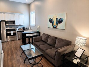 A compact living space with a kitchenette, a brown sofa, a glass coffee table, and a painting of a cow on the wall. - King-bed Historic loft near East Nashville with parking (Nashville)