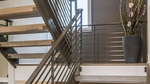 Una escalera moderna con barandas de metal y escalones de madera, que conduce a una habitación con un ventanal grande y una planta en maceta.