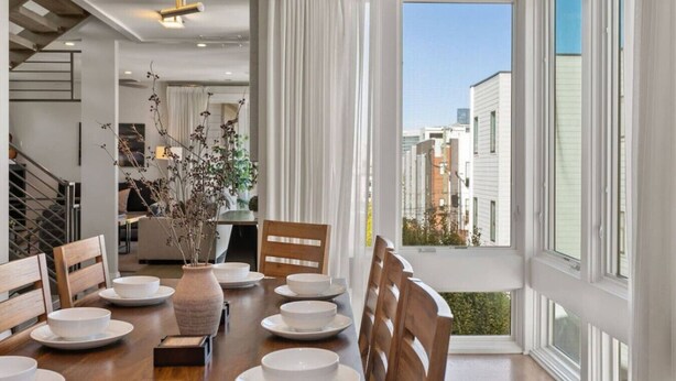 Comedor con mesa de madera para cuatro, platos blancos y un florero con ramas secas.