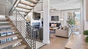 Un interior moderno con una escalera de madera, una sala de estar con sofá y mesa de centro, y una cocina con televisor y armarios.