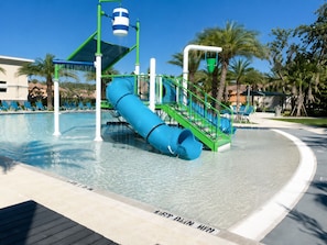 Un parc aquatique avec un toboggan bleu, une fontaine et un espace piscine équipé de chaises longues.