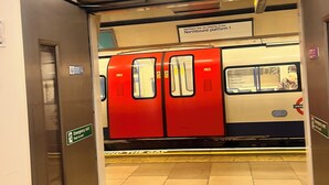 Una stazione della metropolitana con un treno rosso, segnali di uscita di sicurezza e un avviso di bordo piattaforma.