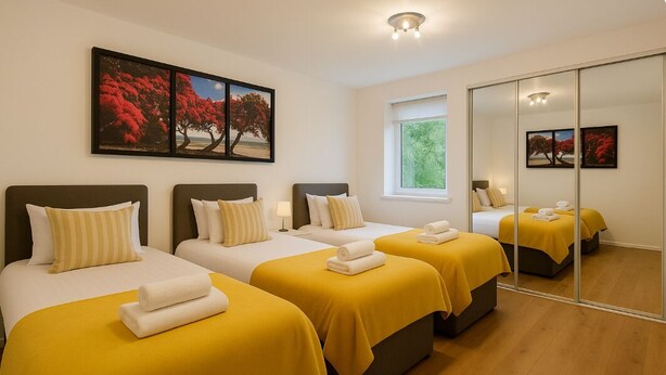 Una camera d'albergo con tre letti, ognuno con una coperta gialla e cuscini bianchi, un'ampia finestra e uno specchio che riflette la stanza.