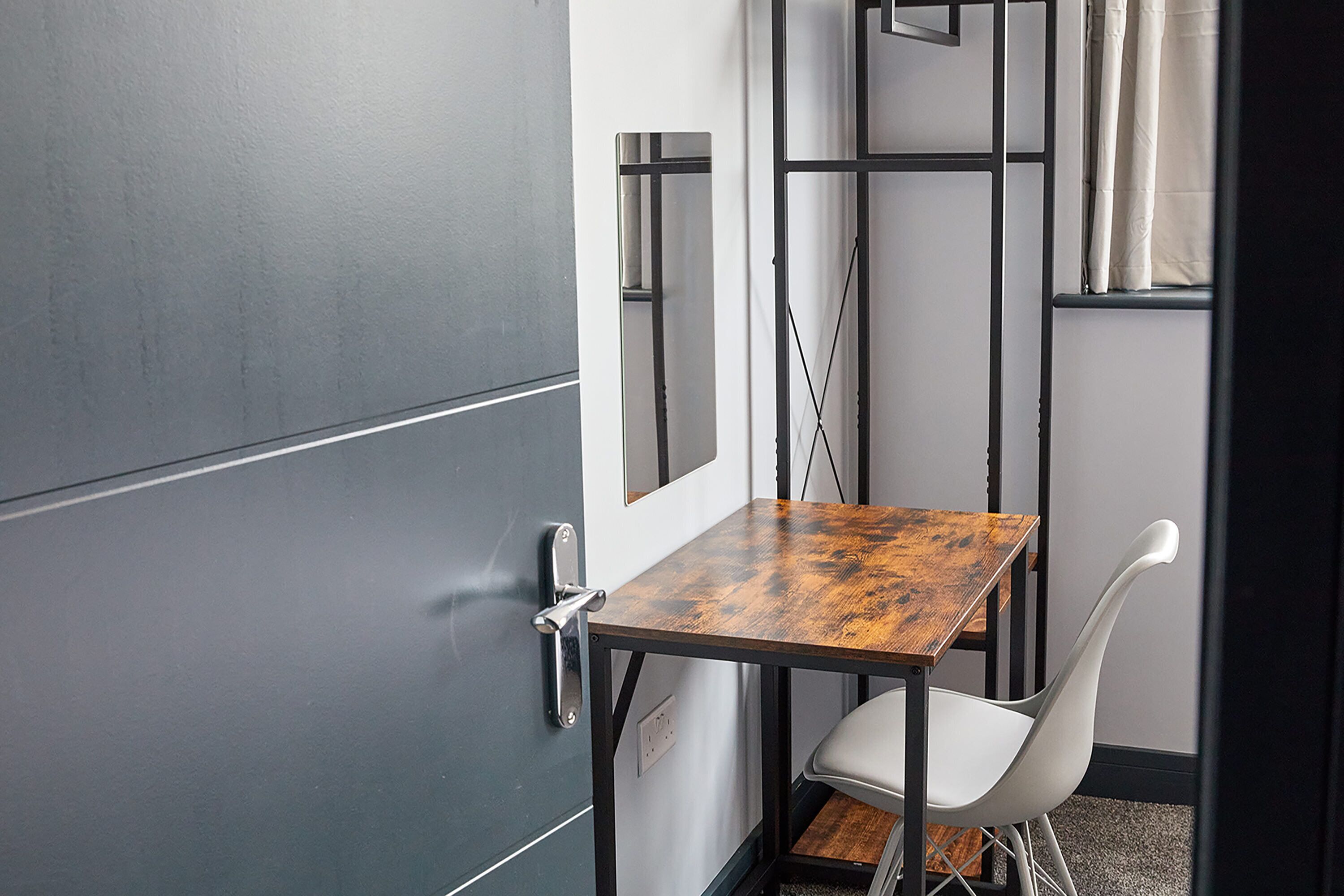 A small room with a dark door, a wooden table, a white chair, and a mirror on the wall.