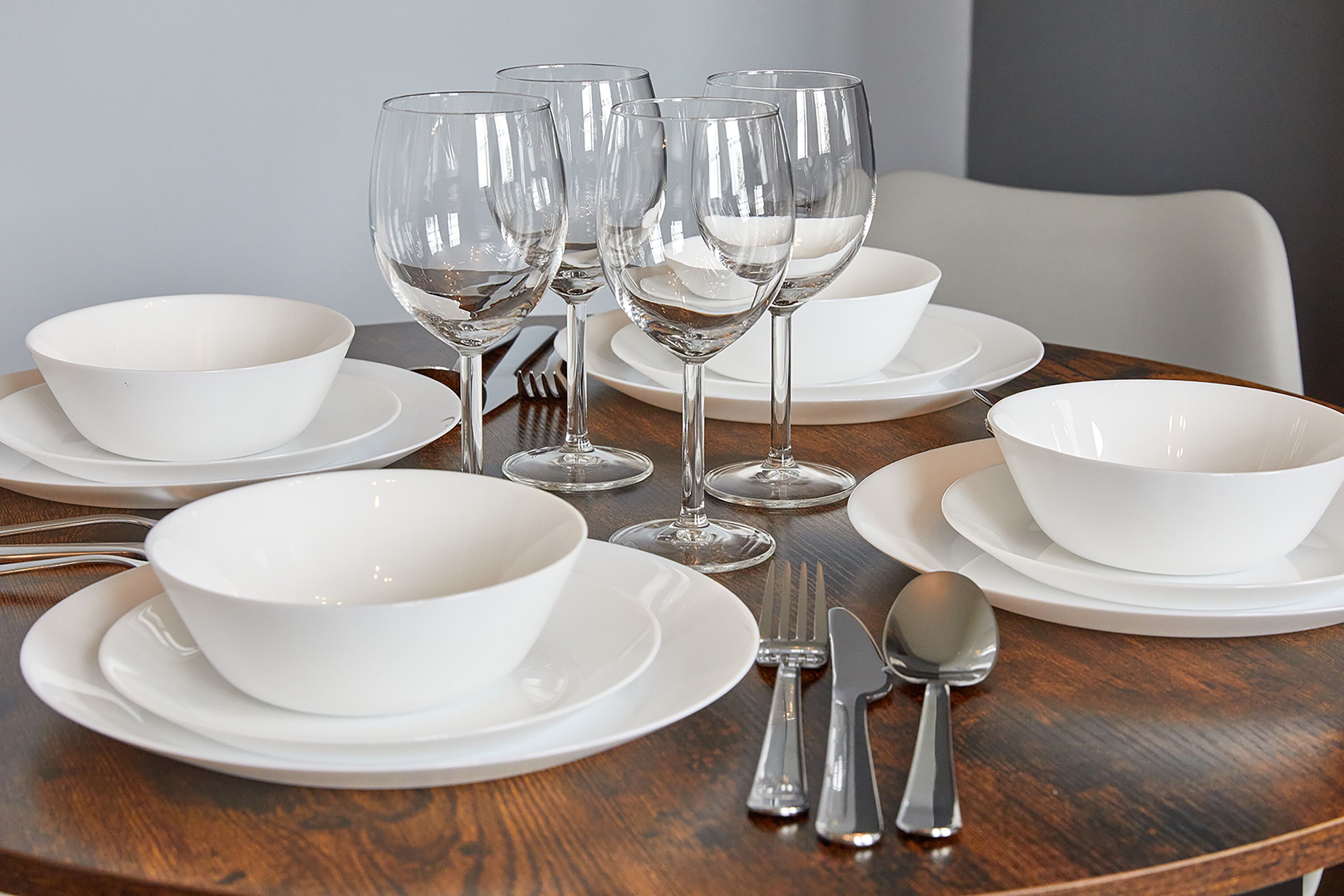 Una mesa de comedor dispuesta con fuentes, platos y cubiertos blancos, incluyendo copas de vino y una cuchara.