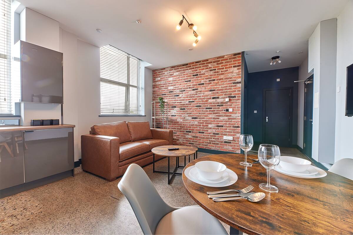 A modern kitchen with a brick wall, a dining area with a wooden table set for two, and a leather sofa.