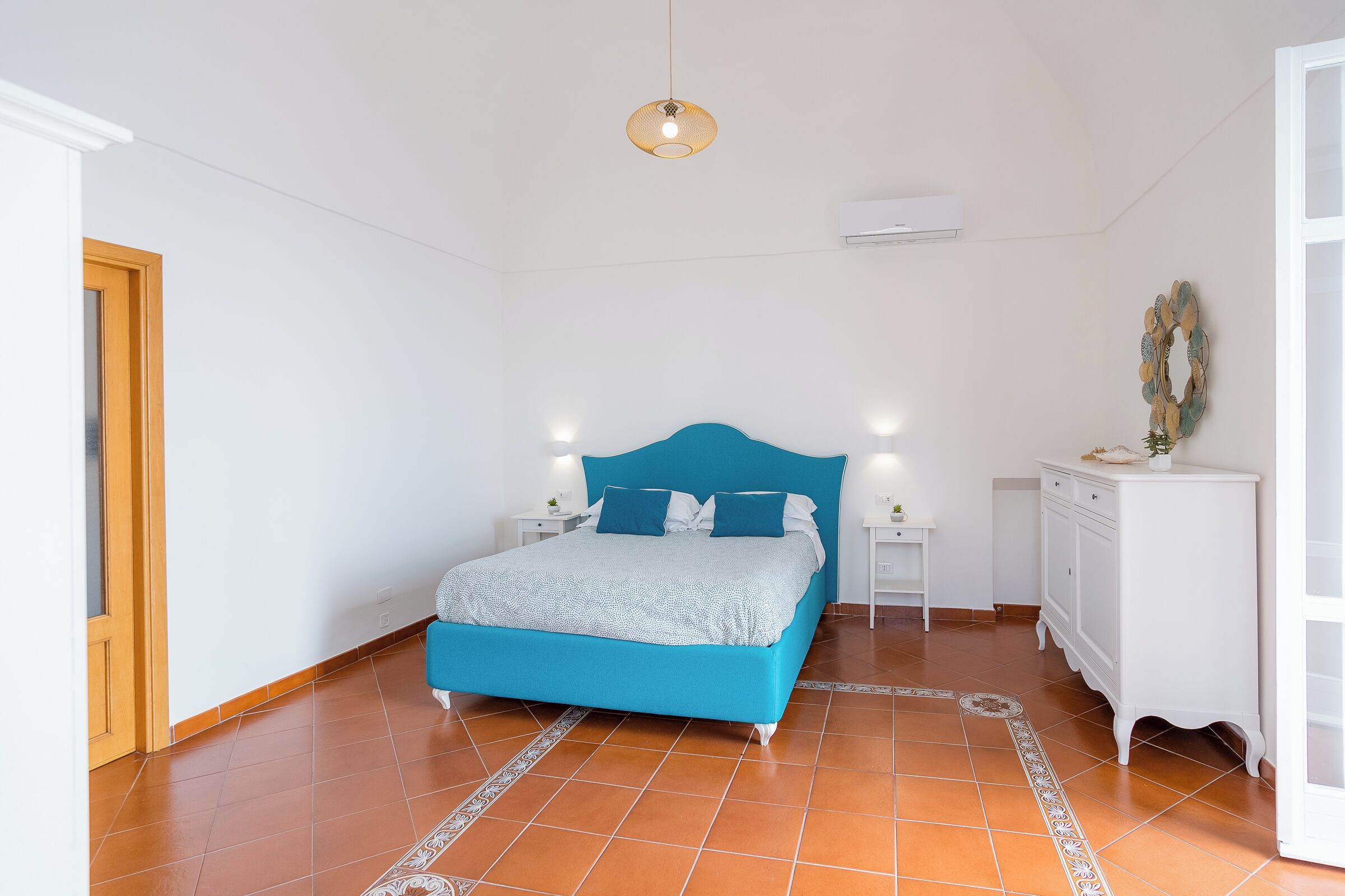 A bedroom with a blue bed, white walls, a wooden door, a white dresser, and a patterned rug.