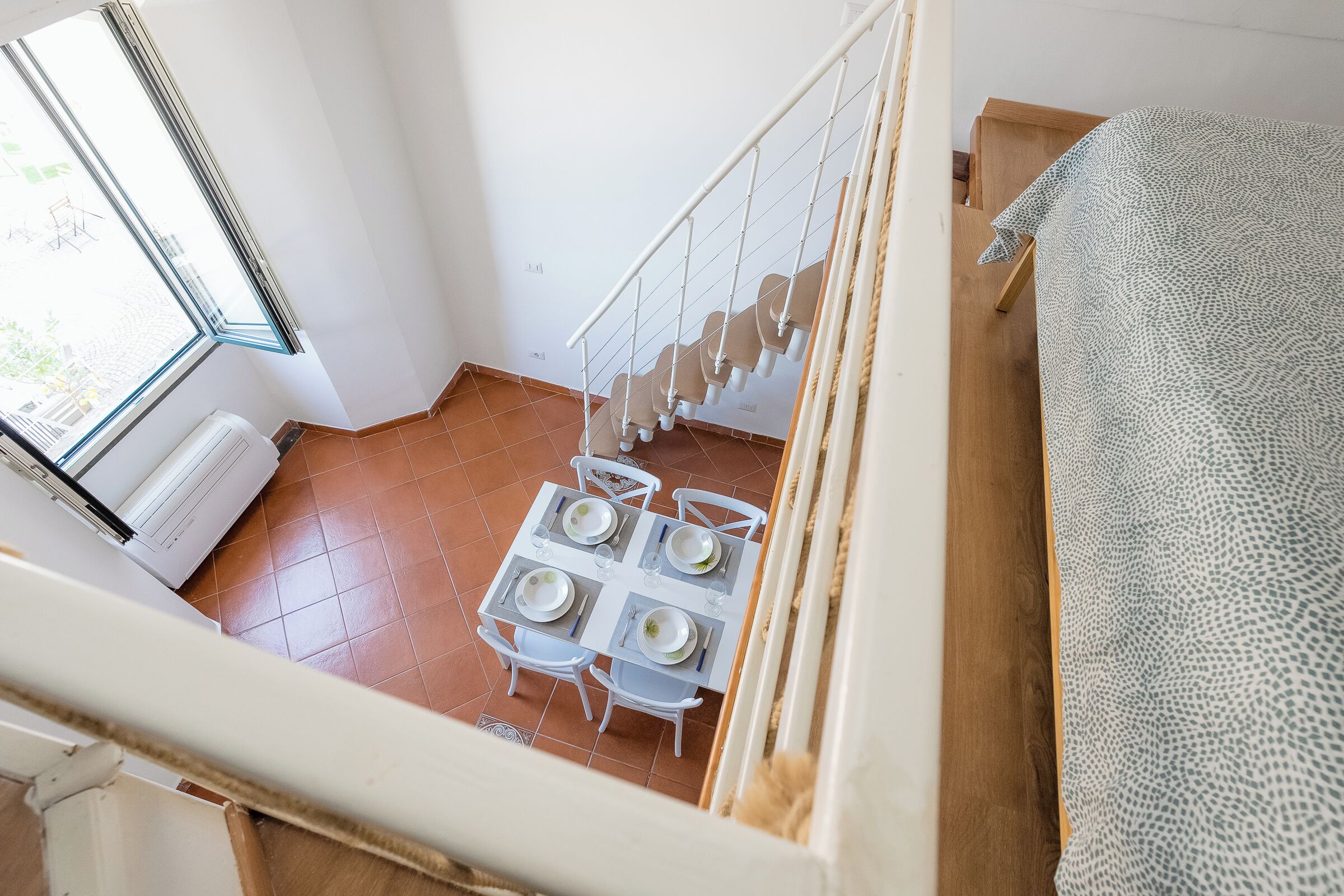 Un comedor con una mesa preparada para cuatro personas, una escalera y un ventanal con persianas.