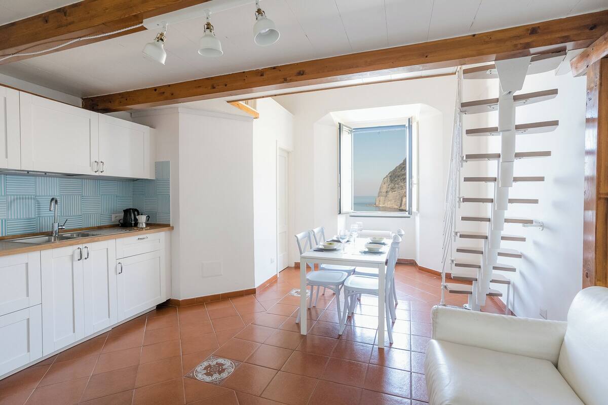 Une cuisine spacieuse avec des meubles blancs et un sol carrelé, un coin repas avec une table et des chaises blanches, et une vue sur la mer par une grande fenêtre.