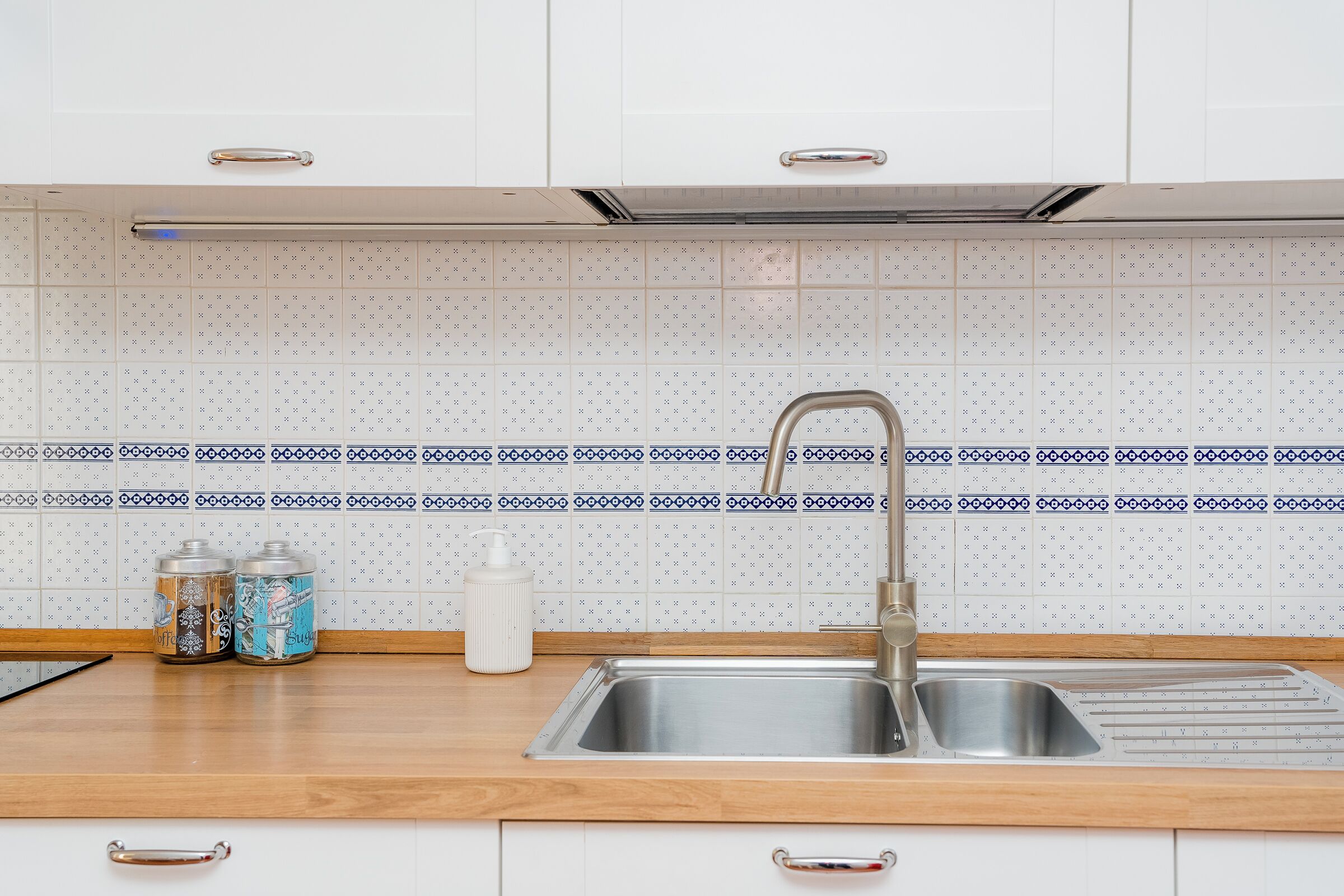 A kitchen with a stainless steel sink, wooden countertop, and patterned backsplash.