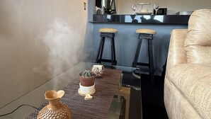 En moderne stue med et glastæppe, en fjernbetjening og en dekorativ vase. I baggrunden er der et køkkenområde med en barstol og en sofa.