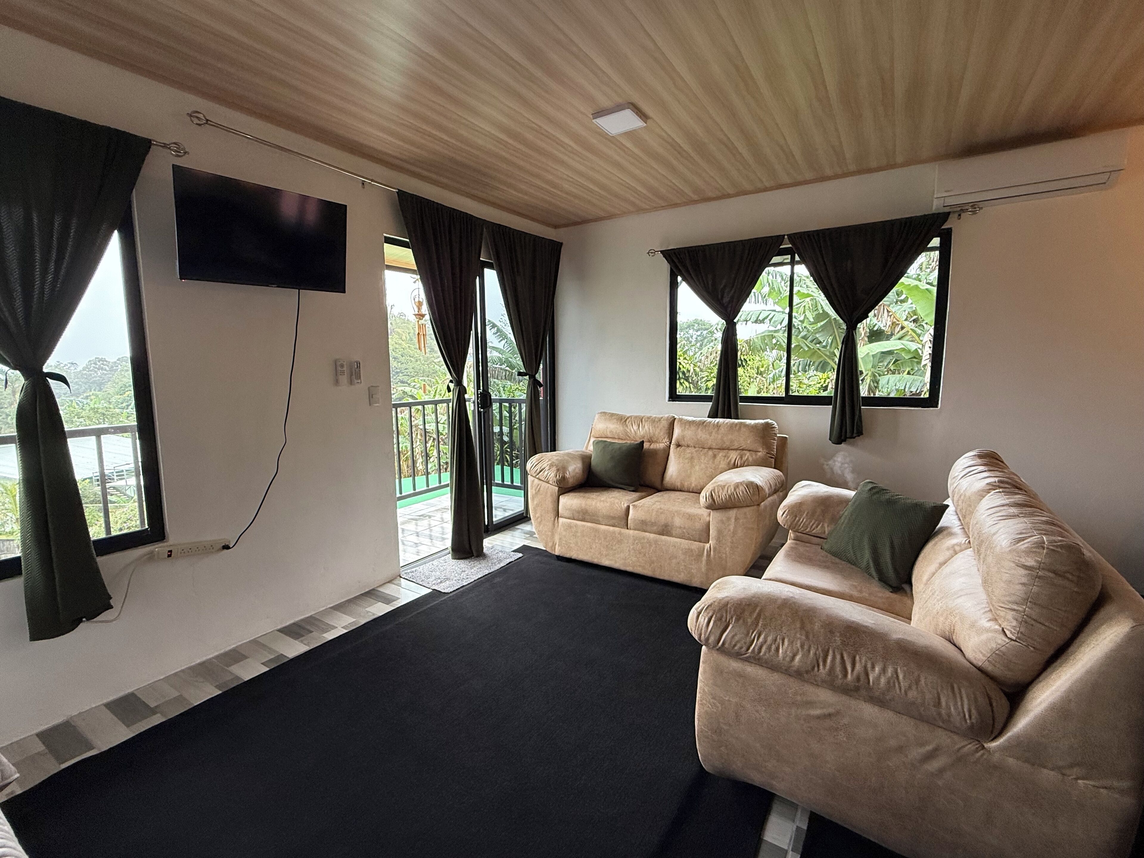 Een woonkamer met een beige bank, een flatscreen-tv aan de muur en een balkon met gordijnen.
