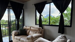En rummelig stue med en beige sofa, en mindre lænestol og udsigt til frodig vegetation og et bjerg gennem store vinduer.