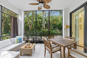 A room with a large sliding glass door, a ceiling fan, a dining table with chairs, a sofa, and a small side table. - Serene Resort Lifestyle Condo Near Beach & 5th Ave (Naples)