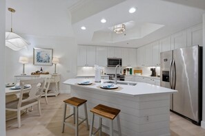 A modern kitchen with white cabinetry, a central island with stools, a stainless steel refrigerator, and recessed ceiling lighting. - Serene Resort Lifestyle Condo Near Beach & 5th Ave (Naples)