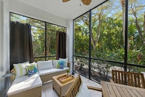 A sunlit room with a large window, a sofa with cushions, a wooden table, and a chair. - Serene Resort Lifestyle Condo Near Beach & 5th Ave (Naples)