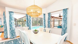 Un comedor con una mesa blanca, sillas blancas, una lámpara de techo de mimbre y cortinas con estampado azul.