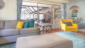 Una sala de estar con un sofá, un reposapiés y un sillón amarillo. Se observa un comedor con una mesa y sillas a través de una puerta de vidrio.