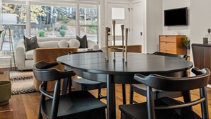 Un comedor moderno con una mesa redonda y sillas negras, un sofá con cojines, y un televisor fijado en la pared.