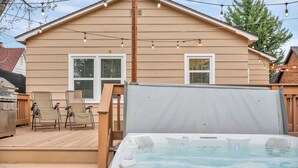 Ein Whirlpool auf einer Holzterrasse mit Lichterketten, einer überdachten Terrasse und einem Haus mit zwei Fenstern und einem Schornstein.