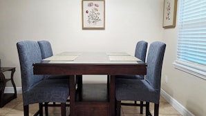A dining table with four chairs, a framed floral artwork on the wall, and a window with blinds. - Toria Suites Condo 924 (Lakeland)