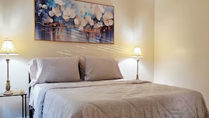 A bedroom with a bed, two bedside lamps, a large floral painting, a bench, and a striped rug. - Toria Suites Condo 924 (Lakeland)
