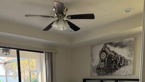 Ein Schlafzimmer mit einem Bett, einem Deckenventilator, einem Gemälde und Blick ins Freie.
