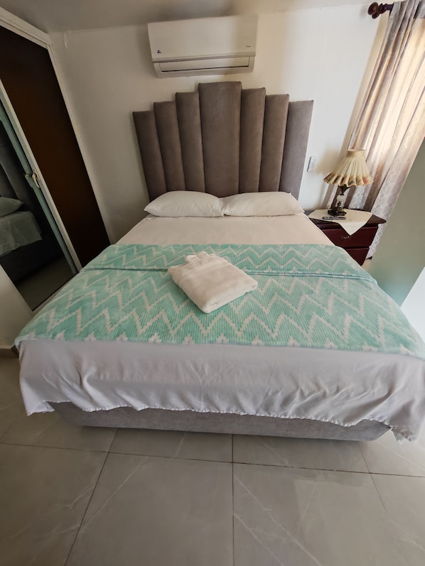A neatly made bed with a patterned bedspread, a bedside table with a lamp, and an air conditioning unit on the wall.