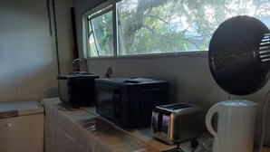 Eine Küchenzeile mit Mikrowelle, Toaster und Kaffeemaschine auf einem Tisch, ein kleiner Kühlschrank und ein Fenster mit Blick auf Bäume.