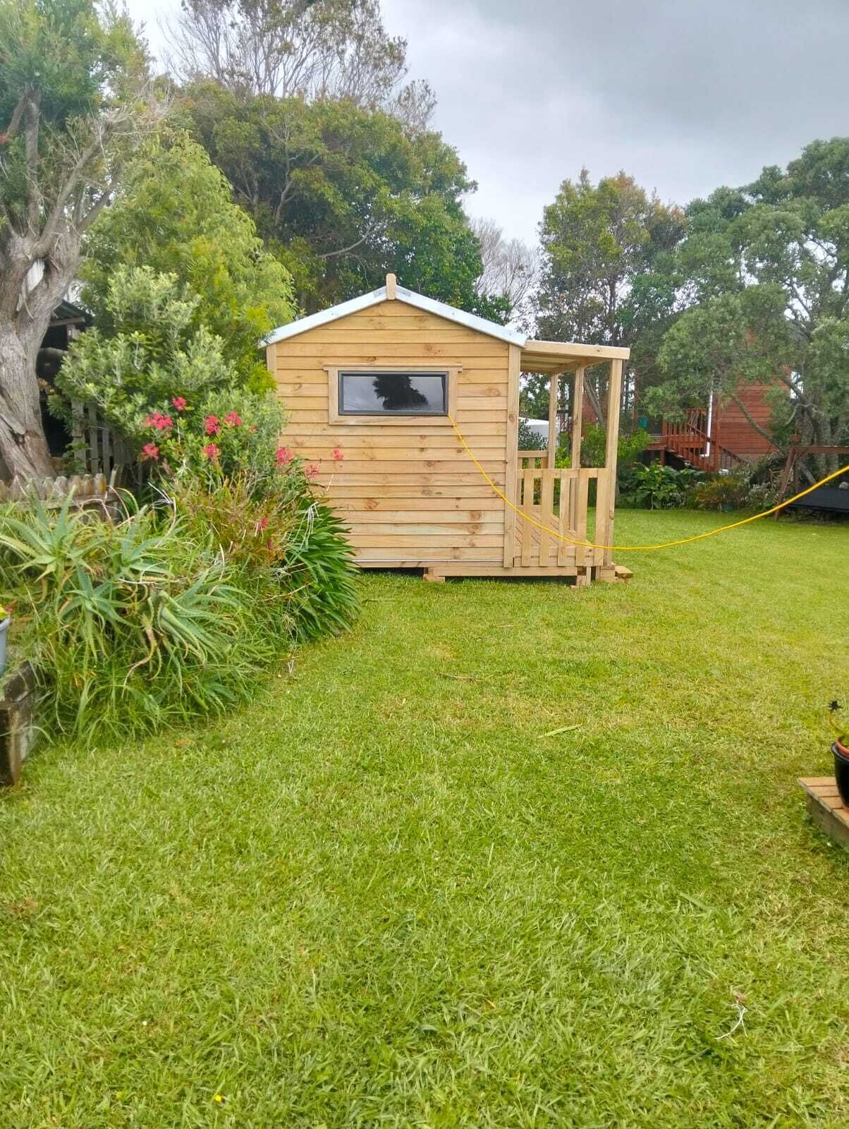 Une petite cabane en bois, dotée d’une fenêtre et d’une terrasse, entourée d’une végétation luxuriante et d’arbres.