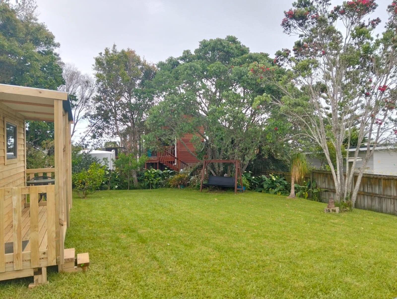 Una casita de juegos de madera en un patio trasero con un columpio, un cobertizo pequeño y varios árboles y arbustos.