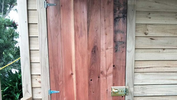 Eine Holztür mit Metallriegel und Klinke, eingebaut in eine Holzkonstruktion mit Holzgeländer und Fußmatte auf der Veranda.