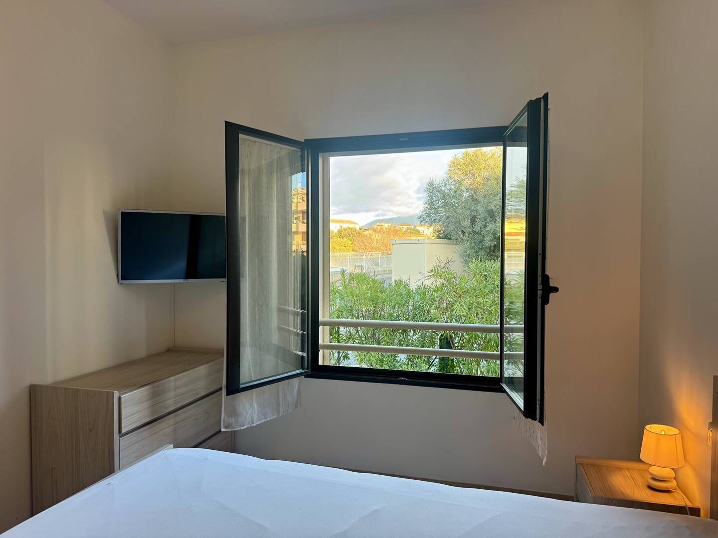 Een hotelkamer met een bed, een aan de muur bevestigde televisie, een raam met een open gordijn en een nachtkastje met een lamp.