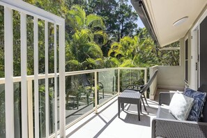 Ein Balkon mit Glastrennwand, Korbmöbeln und üppiger Bepflanzung im Hintergrund.