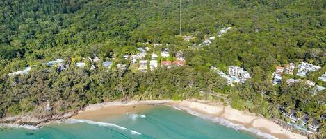 A coastal area with a beach, dense forest, and a white structure on a hill.