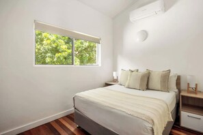 Ein Schlafzimmer mit einem Bett, einem Nachttisch, einem Fenster mit Blick auf Bäume und einer Wand-Klimaanlage.
