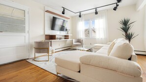 Una sala de estar moderna con un sofá, una mesa de centro y un televisor. Hay una planta y una ventana con cortinas.