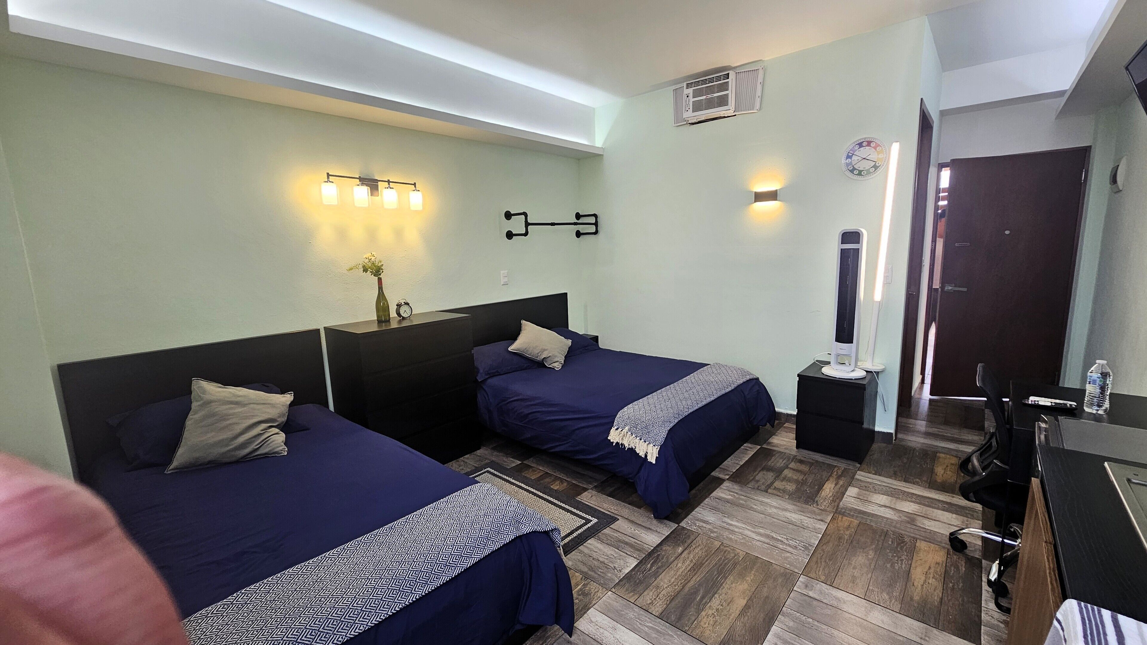 A hotel room with two beds, a desk, a chair, a nightstand, a mirror, a clock, and a vase with flowers.