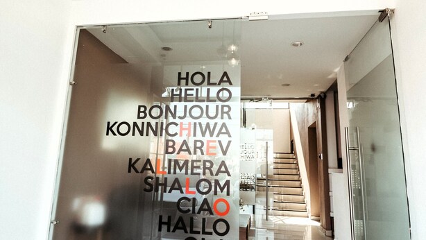 A modern hotel corridor with a glass wall displaying multilingual greetings, a staircase, and a clean, tiled floor. - A30 Plaza Acequia Suite Black Plus   (Cuautitlán Izcalli)