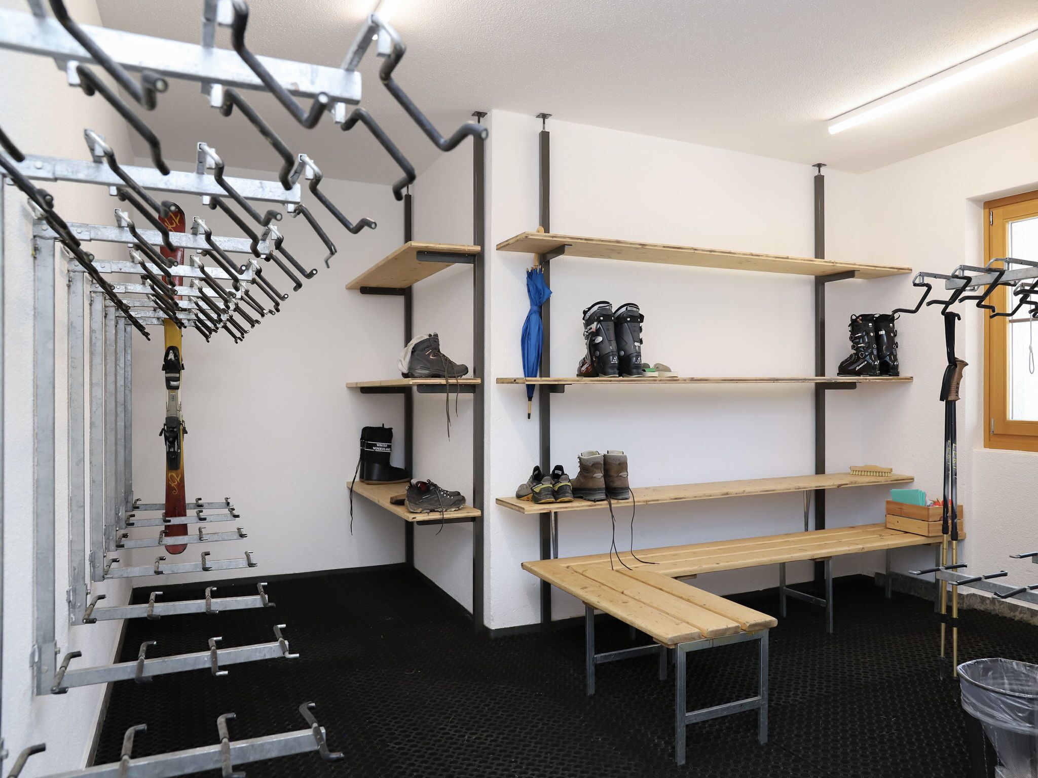 Un depósito con estanterías metálicas para esquíes y botas, repisas de madera para zapatos, y un banco.