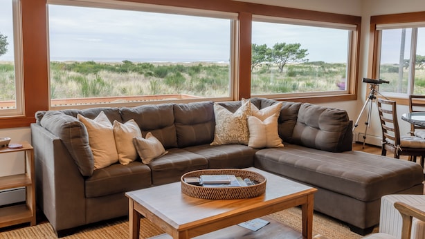 A modern living room with a sectional sofa, a coffee table, and large windows offering a view of the beach and dunes. - Seaside Oceanfront on the Prom | 5BR, Sweeping Views, and Direct Access to the Seaside Promenade (Seaside)