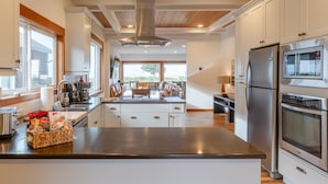 A modern kitchen with stainless steel appliances, a central island, and a dining area with a view. - Seaside Oceanfront on the Prom | 5BR, Sweeping Views, and Direct Access to the Seaside Promenade (Seaside)