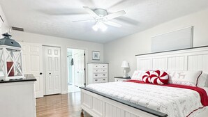 Quarto com cama branca, piso de madeira, ventilador de teto e paredes brancas.