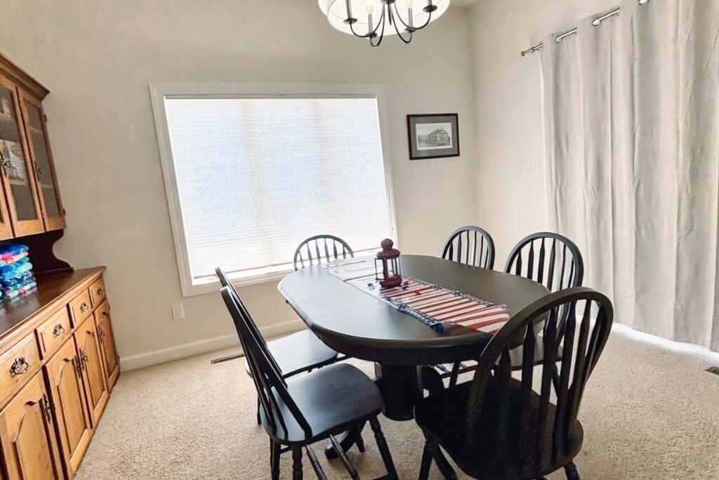A dining room with a table, chairs, a chandelier, a window with blinds, a cabinet, and a picture on the wall.