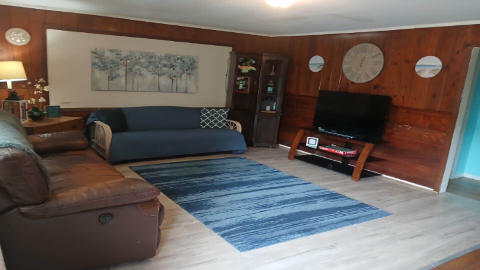 Een woonkamer met een blauwe bank, een bruine leren fauteuil, een flatscreen televisie op een houten standaard, een blauw vloerkleed en een wandklok.